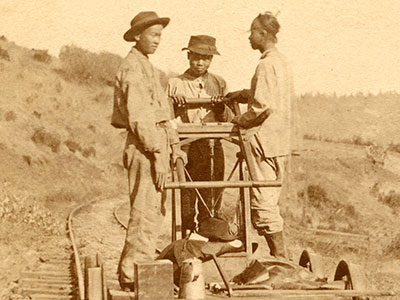 A vintage photograph of four men standing on a handcar on a railway track in a rural landscape. Three men are on the handcar, wearing hats and work clothes, while one man in a suit and top hat stands on the ground beside the track.